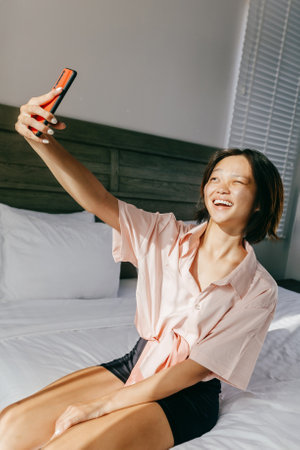 Asian young adult female smiling and taking a selfie on bed in bright room.