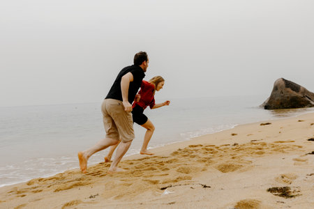 Young caucasian couple running and laughing on sandy beach by the ocean.の写真素材