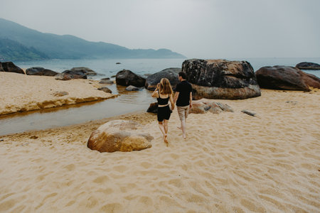 Young caucasian couple walking on sandy beach with boulders and ocean view.の写真素材