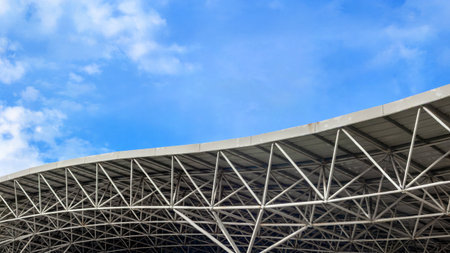 Curved steel roof structure against blue sky with clouds.