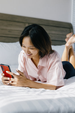 Young Asian female relaxing on bed using smartphone in casual pink shirt.の写真素材