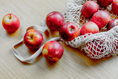 Fresh red apples spilling from reusable mesh bag on wooden surface.の写真素材