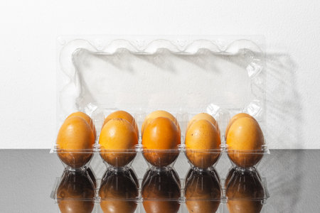 Five brown eggs in a transparent plastic cardton on reflective surface.