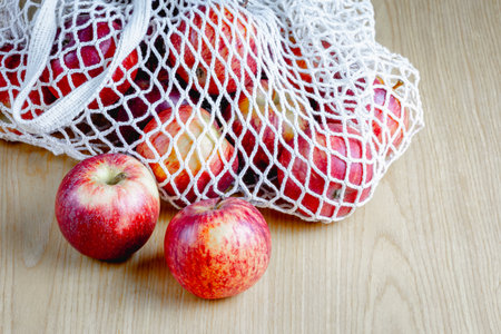 Red apples in mesh bag on wooden table - fresh, ripe, healthy, natural fruit display.の写真素材