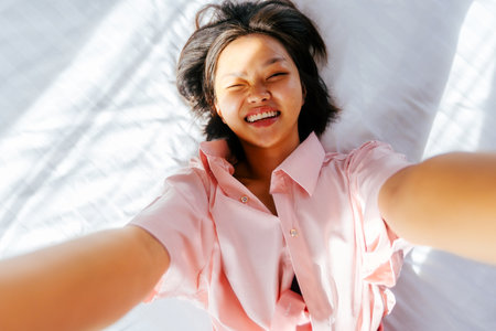 Asian young woman smiling in pink shirt lying on bed in natural light.の写真素材