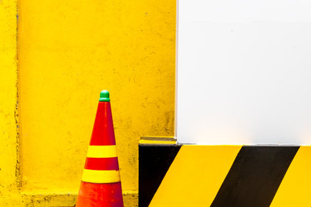 Vibrant traffic cone against yellow wall with black and yellow striped barrier.