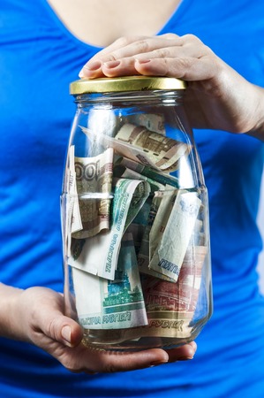 The woman in a blue shirt holding jar with Russian money on a grey background.