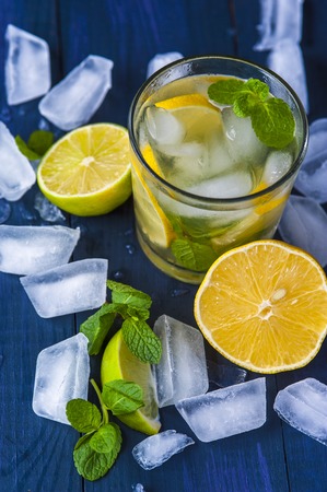 Glass of homemade lemonade with slices of lemon and lime, mint leaves and ice cubes on wooden rustic background. Focus on lemon. Vertical view.の写真素材