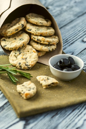 Homemade cookies with cheese, olives and rosemary, cup of olives and rosemary branches on linen napkin on wooden rustic background. Focus on cookies. Vertical view.の写真素材