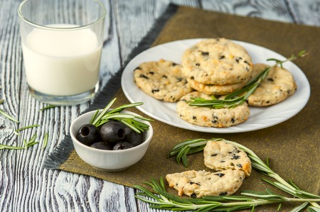Homemade cookies with cheese, olives and rosemary on white plate, cup of olives, rosemary branches on linen napkin and glass of milk on wooden rustic background. Focus on cup. Horizontal view.の写真素材