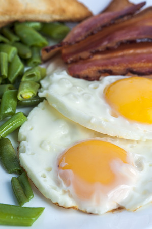 Fried eggs, bacon, green beans and toasts on light background. English breakfast. Vertical view. Focus on eggs.の写真素材