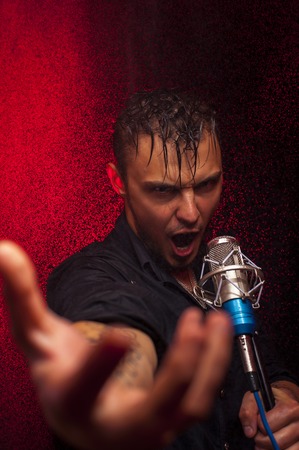 Portrait of young expressive male singer with microphone. Man looking at camera, hand reaching out. Red spotlight background.の写真素材