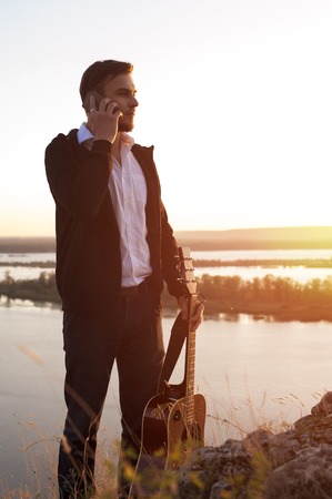 Young attractive man in casual cloth staying on the beach, talking on the phone and holding acoustic guitar, sunset.の写真素材