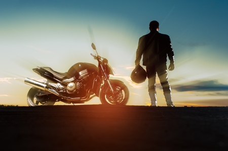 Silhouette of male biker in leather outfit standing next to bike with helmet at sunset timeの写真素材
