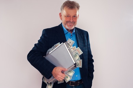 Portrait of smiling caucasian senior man holding metal briefcase with dollar bills. Financial...
