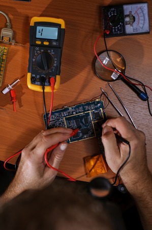 Electrician checking circuit computer board with multimeter. Magnifier, tester, tweezers on table....