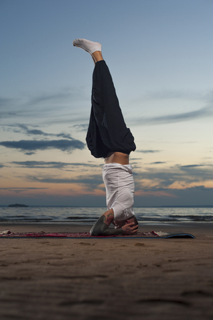 Full length shot young tattoo man practicing yoga on the beach at sunset. Headstand or shoulderstand exercise.の写真素材