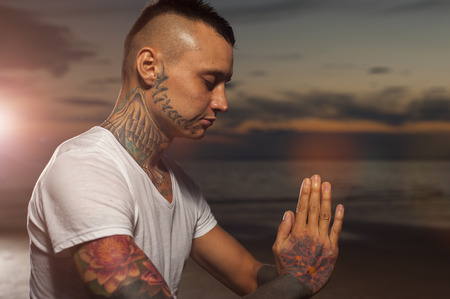 Young attractive man with tattoos on the body practicing yoga and meditating on the beach at sunset.の写真素材