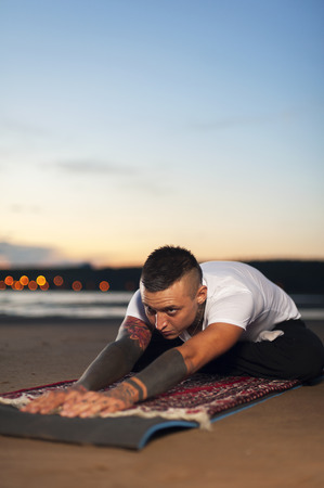 Young flexible man with tattoos on the body doing yoga on the beach at sunsetの写真素材