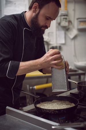 Young bearded male chef grating cheese on panの写真素材