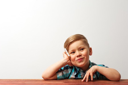 Funny smiling child playing with smartphone. Cute caucasian boy sitting at the table and talking on...