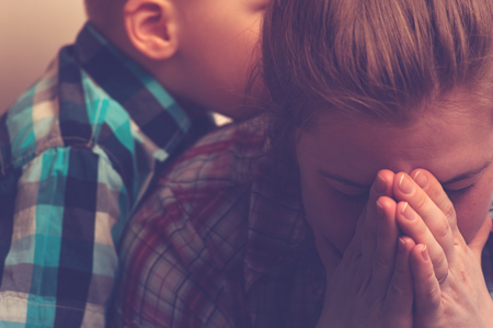 Crying unhappy woman covering her face with hands while little child standing next to her. Tired and depressed mother with kid at home. Family relations problems.の写真素材