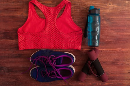 Sports and fitness equipment: dumbbells, water bottle, sports shoes and red bra on wooden...