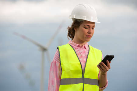 Portrait of a beautiful positive woman architect in a green vest shirt with a smartphone in hand against the backdrop of a windmillの写真素材