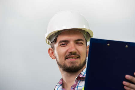 Portrait of a serious ambitious engineer wearing white hard hat against the backdrop of sky.