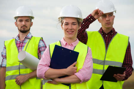 Concept of team work. Three engineers in white helmets and green vests standing with plans against...