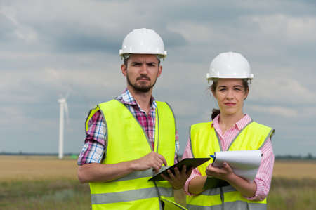 Concept of alternative energy and windmills. Two engineers in white helmets and green vests with working plans on the background of windmills farmの写真素材
