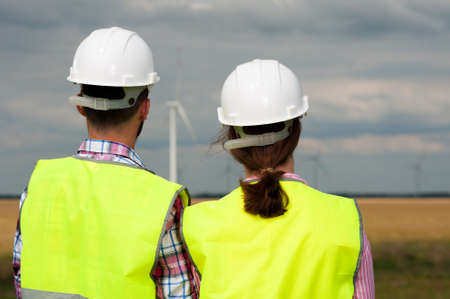 Concept of alternative energy and windmills. Rear view of two engineers in white helmets and green vests looking somewhere into the distanceの写真素材