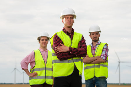Concept of engineers and windmills. Three engineers in white helmets and green vests standing with...
