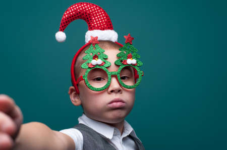 Sad little boy wearing headband with Santa Claus Hat and funny glasses with Christmas trees posing on green wall. Merry Christmas and Happy New Year child.の写真素材