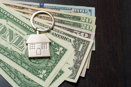 Close-up keychain in the form of house lies in it a pile of dollar bills lying on a black surface....