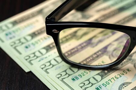 Close-up part of the glasses lie on dollar bills on a black table. Concept of work as an accountant or financier. Concept of stable salary and businessの写真素材