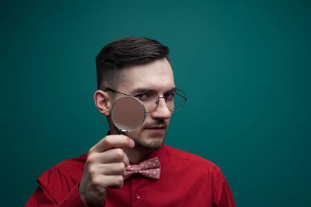 Portrait of a young handsome guy looking through a magnifying glass on a green backgroundの写真素材