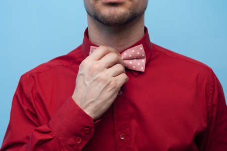 Close-up of the hands of a young man in a red shirt correcting bow-tie against a blue background. The concept of gallantry and styleの写真素材