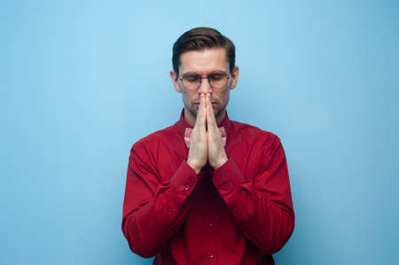 Portrait of a begging young man in glasses and a red shirt with crossed hands at his lipsの写真素材
