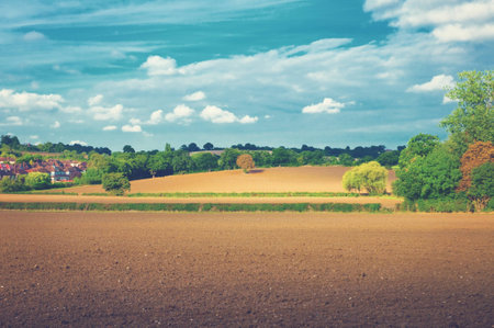 Beautiful afternoon landscape with plowed fields on hills and houses in the background under cloudy sky.の写真素材