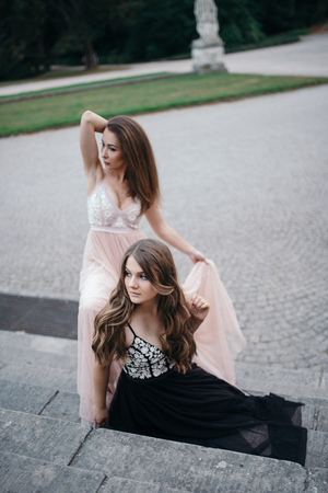 Portrait of two beautiful brunettes in long chiffon dresses standing and sitting at a parkの写真素材