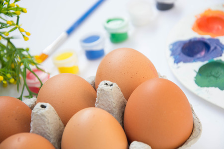 Easter brown chicken eggs in carton container, with plastic flowers and blurred paints, colorful palette on the background, paint equipment on white backgroundの写真素材