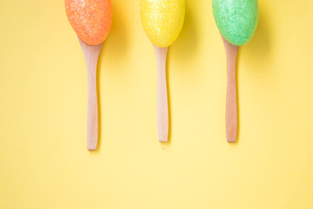 Three Colorful Easter eggs on pale yellow background, egg in wooden spoons, green, orange, yellow, egg hunt decoration, top view - flat layの写真素材