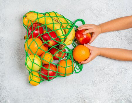 Children's hands put fruit in a string bag with mixed fruits: bananaite, lemons, apples, oranges....