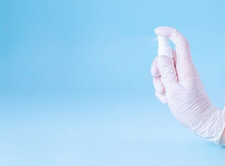 Female hand in a rubber white glove sprays hand antiseptic from a white bottle on a blue background. Banner. Coronovirus, Covid-19, Quarantine, Pandemic. Close-up. Flat lay, copy space.の写真素材