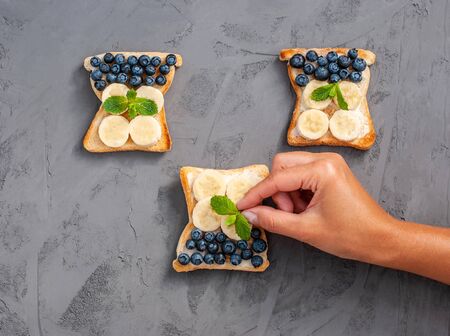 Female hand holds lays a wash sheet on a homemade sandwich with cottage cheese, blueberries, banana, on a dark background, copy space. Flat lay. Healthy snack, proper nutrition. View from above.の写真素材