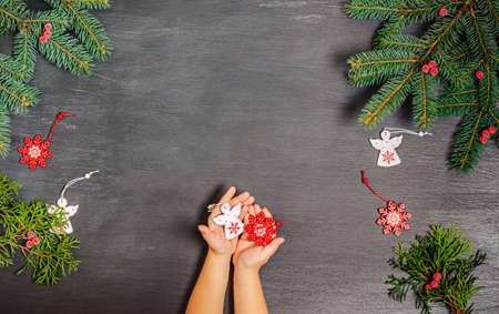 Children's hands hold red and white wooden Christmas toys with juniper branches on a black chalk background. Banner, form for a postcard. Copy space, flat lay. New Year, Christmas, 2021. Top view.の写真素材