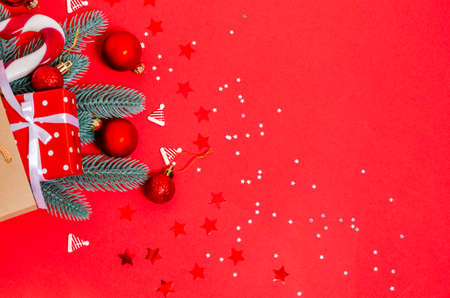 Christmas toys, gift box, fir branches, caramel cane in a paper shopping bag on a red background....