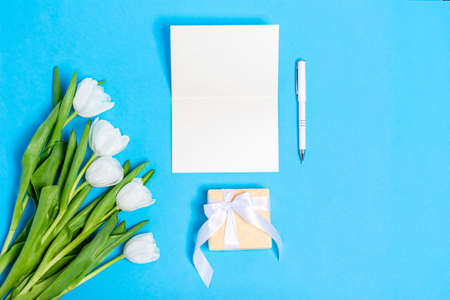 Bouquet of white tulips with gift boxes and blank paper on a blue background. Flat lay, copy space,...