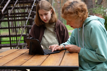 computer awareness of the elderly. a happy teenage girl teaches older 70s grandmothers to use a laptop at home. social media and modern technologies: a pensioner studies in online security.の写真素材